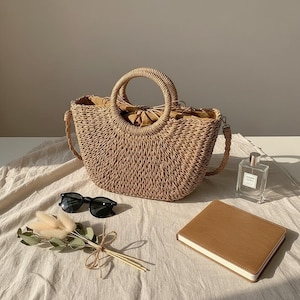May include: A woven straw handbag with a circular handle and a shoulder strap. The bag is tan and sits on a white cloth, next to black sunglasses, a brown notebook, a perfume bottle, and dried flowers.