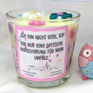 Könnte beinhalten: Eine Glas-Kerze mit gelbem Wachs und floralen Verzierungen. Die Kerze hat ein Etikett mit deutschem Text und einer Berliner Skyline. Rechts befindet sich eine Eulenfigur.