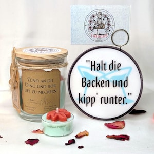 Könnte beinhalten: Ein Glas-Kerzenglas mit Holzdeckel und dem Text "Zünd an dit Ding und hör uff zu meckern." Eine kleine, runde Kerze mit Blumenform und dem Text "Patchouli Paradies." Ein Schild mit dem Text "Halt die Backen und kipp' runter."