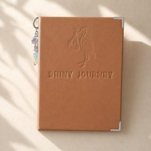 May include: A tan leather journal featuring the embossed words "SHINY JOURNEY" and a stork illustration. The journal has silver corner protectors and a charm with colourful beads. A lovely keepsake for recording memories.