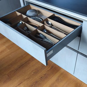 Può includere: Cassetto della cucina aperto con un organizer per posate in legno. Il cassetto contiene vari utensili in acciaio inossidabile, tra cui cucchiai, forchette e utensili da portata. Il cassetto è grigio e poggia su un pavimento in legno.