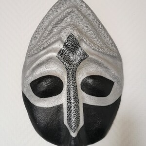 Puede incluir: Una máscara decorativa con una combinación de colores plateado y negro. La parte superior es plateada con un patrón texturizado, mientras que la sección inferior es negra. Un diseño plateado en forma de cruz con un centro texturizado está centrado en la máscara.