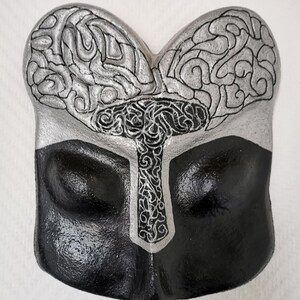 Könnte beinhalten: Eine zweifarbige Maske mit einem gehirnähnlichen Design. Die obere Hälfte ist silberfarben mit schwarzen Liniendetails, die einem Gehirn ähneln. Die untere Hälfte ist schwarz. In der Mitte befindet sich eine dekorative silberne kreuzartige Form.