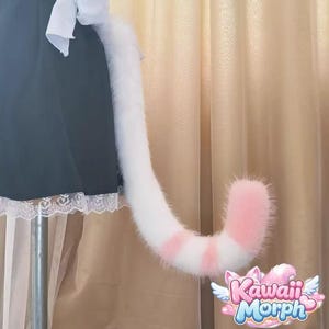 May include: A fluffy white and pink cat tail with pink stripes. The tail is attached to a black skirt with white lace trim. The tail has a curved shape and is made of soft, faux fur. The logo "Kawaii Morph" is visible.