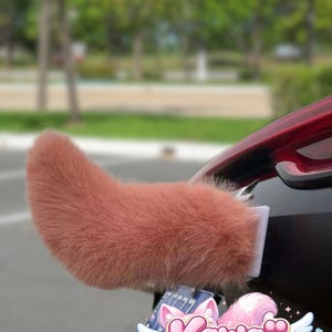 May include: A fluffy, reddish-brown car tail accessory, shaped like an animal tail, attached to a black vehicle. The accessory is mounted with a white bracket. The logo "Kawaii Morph" is visible below the tail.