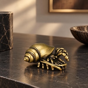 May include: A small, metallic gold-toned figurine of a hermit crab. The sculpture features a detailed spiral shell and articulated legs, showcasing a weathered, antique-style finish with dark highlights in the crevices of the metal surface.
