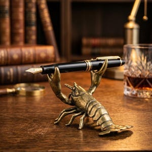 May include: A black fountain pen with gold detailing is displayed on a brass lobster pen holder. The pen holder is on a wooden desk, with antique books and a glass of amber liquid in the background.