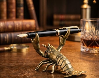 May include: A black fountain pen with gold accents rests on a brass lobster pen holder. The pen holder is detailed and sits on a wooden desk, with antique books and a glass of amber liquid in the background.