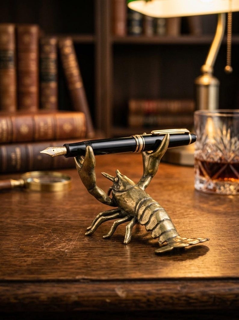 May include: A black fountain pen with gold accents rests on a brass lobster pen holder. The pen holder is detailed and sits on a wooden desk, with antique books and a glass of amber liquid in the background.