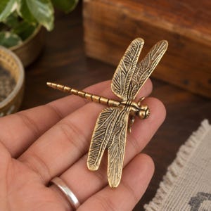 May include: A metallic gold-toned dragonfly figurine featuring intricate, raised wing patterns and a segmented body. The insect has a textured surface with dark antiqued detailing in the grooves, highlighting the veins of the wings and the body segments.