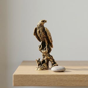 May include: A detailed brass eagle sculpture perched on a tree branch. The eagle is depicted with intricate feather detailing and a focused gaze. A small, smooth stone rests near the base of the sculpture, which is displayed on a wooden surface.