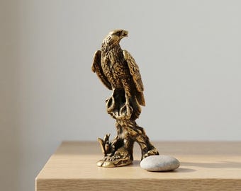 May include: A detailed brass eagle sculpture perched on a tree branch. The eagle is depicted with intricate feather detailing and a focused gaze. A small, smooth stone rests near the base of the sculpture, which is displayed on a wooden surface.