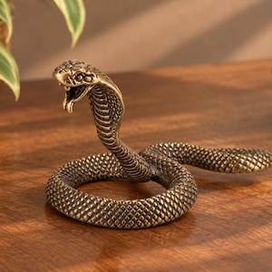 May include: A metallic bronze-toned cobra figurine with a coiled body and an upright, flared hood. The snake features detailed scale textures across its entire surface, with its mouth open to reveal fangs and a forked tongue in a striking pose.