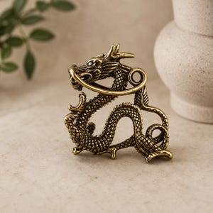 May include: A brass-coloured metal dragon figurine featuring intricate scale detailing, a curved body, and a prominent head with horns. The dragon is depicted in a coiled, serpentine pose with its tail curled at the base and its head turned forward.