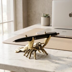 May include: A gold-colored lobster pen holder with a black pen resting in its claws. The pen holder is on a marble desk, with a laptop and small potted plant in the background. The lobster is a decorative desk accessory.