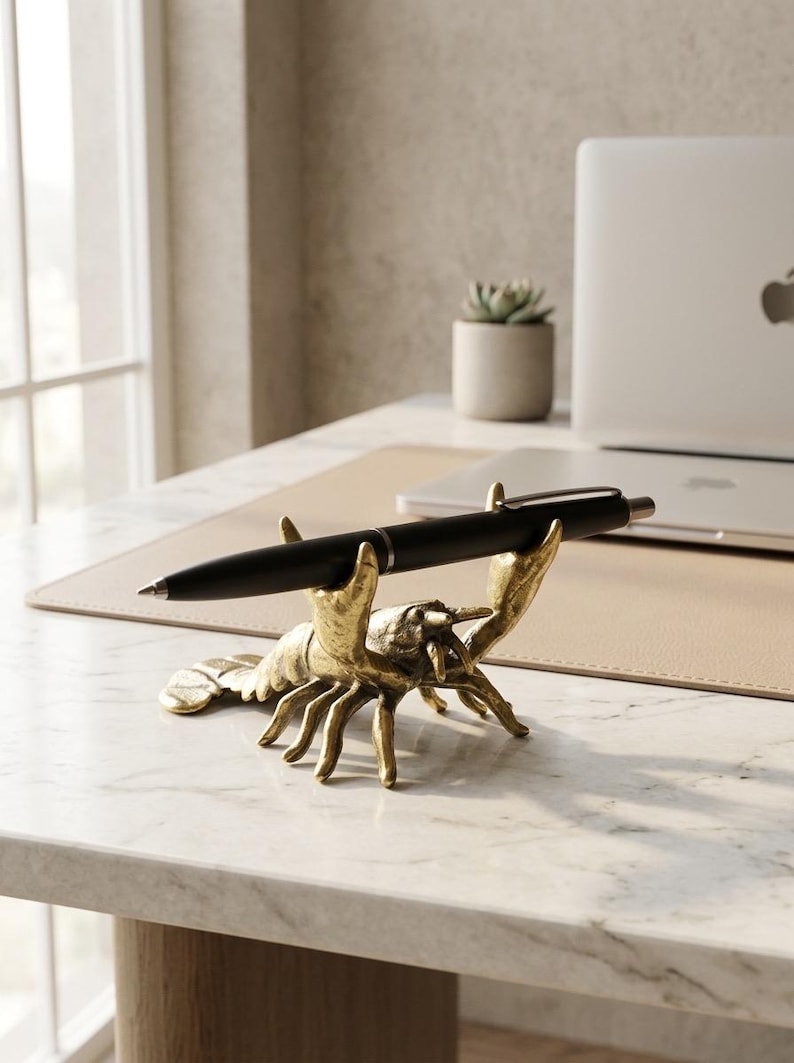 May include: A gold-colored lobster pen holder with a black pen resting in its claws. The pen holder is on a marble desk, with a laptop and small potted plant in the background. The lobster is a decorative desk accessory.