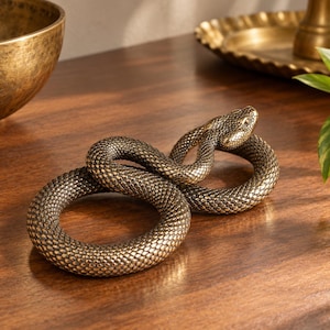 May include: A coiled snake figurine crafted from a metallic bronze-toned material. The sculpture features a highly detailed, textured scale pattern across its entire body, with the head slightly raised and turned to the side in a realistic pose.