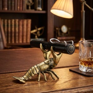 May include: A brass-colored pen holder shaped like a lobster, holding a black pen. The pen holder sits on a wooden desk, with a lamp and a glass of amber liquid in the background. The pen has a silver clip.