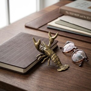 May include: A bronze lobster figurine rests on a brown notebook with a herringbone pattern. Tortoiseshell eyeglasses and a book titled "The Old Man and the Sea" are also on the wooden surface. The scene evokes a nautical or coastal theme.