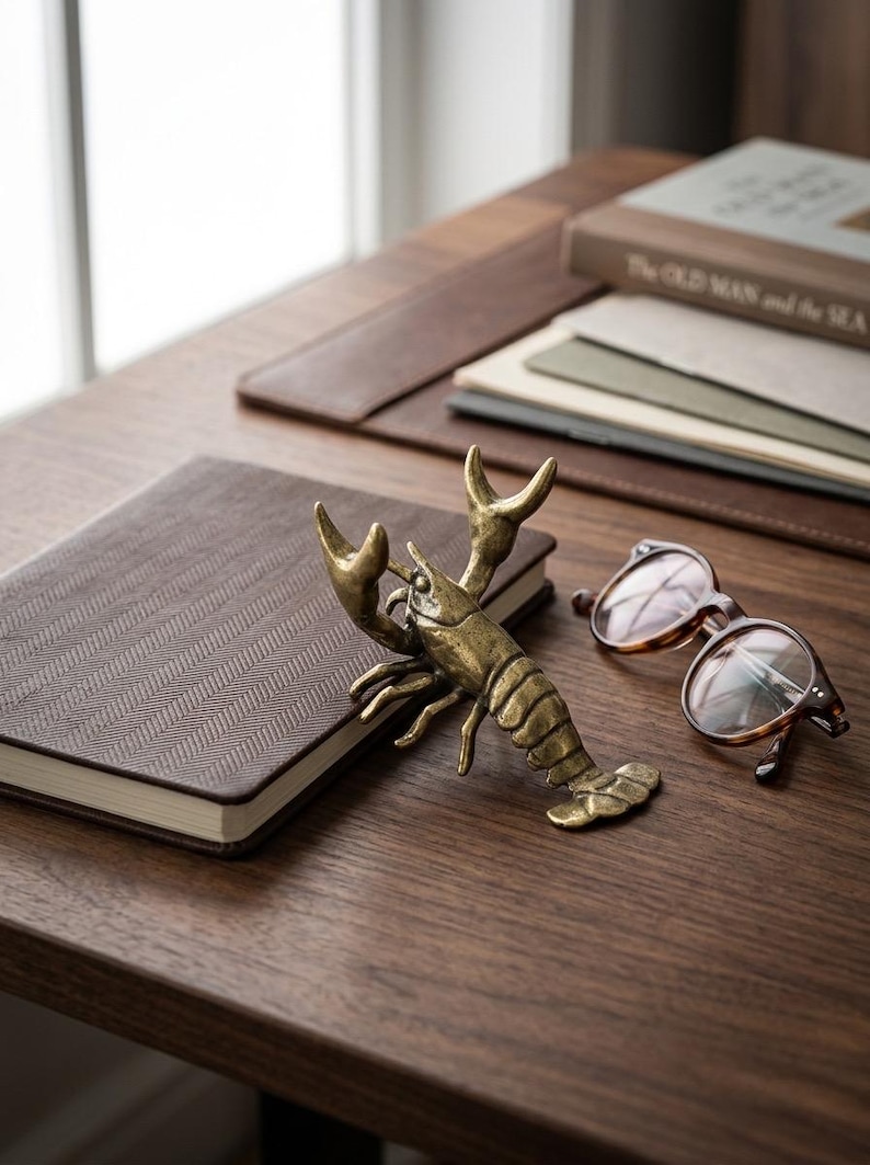 May include: A bronze lobster figurine rests on a brown notebook with a herringbone pattern. Tortoiseshell eyeglasses and a book titled "The Old Man and the Sea" are also on the wooden surface. The scene evokes a nautical or coastal theme.