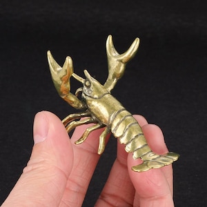 May include: A brass-colored lobster figurine with detailed claws, legs, and segmented body. The lobster is a decorative item, possibly a paperweight or small sculpture. The figurine is held in a hand against a dark background.