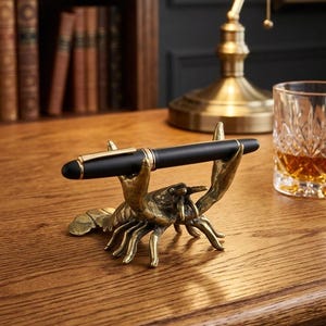 May include: A black pen with gold accents rests in a brass lobster pen holder. The pen holder is on a wooden desk, with a lamp and a glass of amber liquid in the background. The pen holder is a unique desk accessory.
