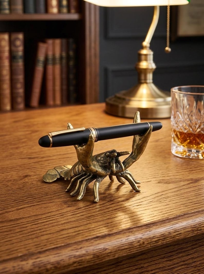 May include: A black pen with gold accents rests in a brass lobster pen holder. The pen holder is on a wooden desk, with a lamp and a glass of amber liquid in the background. The pen holder is a unique desk accessory.