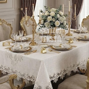 May include: Elegant dining table set for a formal occasion. The table is covered with a white lace-trimmed tablecloth and set with gold-rimmed plates, silverware, and crystal glassware. A floral centerpiece and lit candles add to the ambiance.
