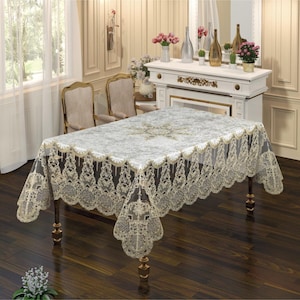 May include: A rectangular tablecloth with a cream-colored lace trim and a floral pattern in the center. The tablecloth is draped over a dark wooden table with ornate legs. The room has a classic, elegant style.