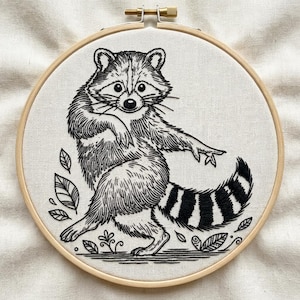 May include: A black and white embroidered raccoon design on white fabric, framed in a wooden hoop. The raccoon is shown in a dynamic pose, with leaves and floral motifs. This handmade artwork would make a unique home decor item.