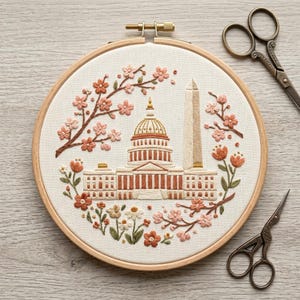 May include: Embroidered artwork in a wooden hoop, depicting the U.S. Capitol and Washington Monument, framed by cherry blossom branches and flowers. The design uses peach, brown, and cream colours. A pair of small scissors is placed beside the hoop.