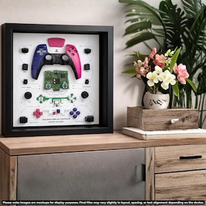 May include: Framed display of a DualSense wireless controller, featuring a disassembled view of its components. The controller's blue and pink halves are visible, along with the circuit board and buttons. The frame is black, and the background is white.
