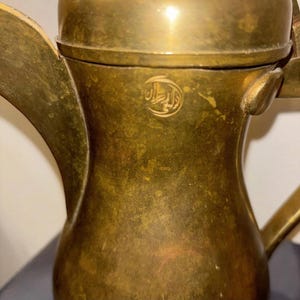 May include: A brass coffee pot with a weathered, tarnished finish. The pot features a curved spout, a lid, and a handle. An engraved circular emblem with Arabic script is visible on the upper body of the vessel, showing signs of age and patina.