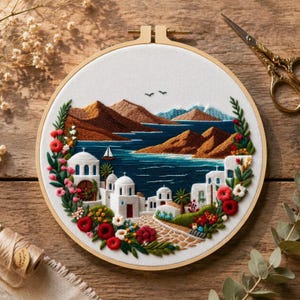 May include: Embroidered hoop art depicting a coastal village with white buildings, blue sea, and brown mountains. The design includes a sailboat, flowers, and greenery, within a wooden embroidery hoop. Gold scissors are visible.