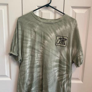 May include: A green tie-dye t-shirt with a black and white telephone graphic on the pocket. The shirt has short sleeves and a crew neck. The shirt is hanging on a black hanger.