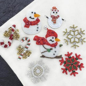 May include: A collection of beaded Christmas ornaments, including snowmen, snowflakes, and candy canes. The snowmen have red scarves and hats, and the snowflakes are in gold, silver, and red. The ornaments are on a white fabric background.