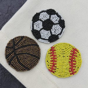 May include: Three beaded sports ball patches: a soccer ball with black and white beads, a basketball with brown and black beads, and a softball with yellow and red beads. The patches are round and sit on a cream-colored fabric.