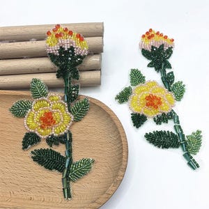 May include: Two beaded flower patches. The flowers have yellow petals with orange centers, green leaves, and stems. The flower buds are yellow, pink, and orange. The patches are on a wooden tray.
