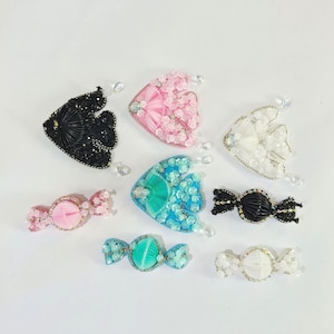 May include: A collection of handmade beaded fish and candy-shaped ornaments in various colors. The ornaments include black, pink, white, and blue fish, and pink, blue, black, and white candy shapes. Each ornament has a clear, teardrop-shaped bead.