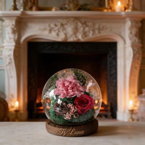 May include: A glass dome terrarium with a wooden base, featuring a pink carnation, a red rose, and greenery. The base is engraved with "To Luna". The terrarium sits on a wooden surface in front of a fireplace.