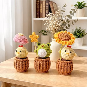 Puede incluir: Tres decoraciones de macetas hechas a mano con ganchillo. Una presenta un pollito con un paraguas rosa, otra una rana con una estrella y la última un pollito con un girasol. Cada una está en una maceta marrón.