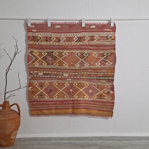 Puede incluir: Textil rectangular tejido con un patrón geométrico en tonos rojos, marrones y amarillos. El textil cuelga de una barra blanca con clips. Un jarrón de terracota con ramas está en primer plano.