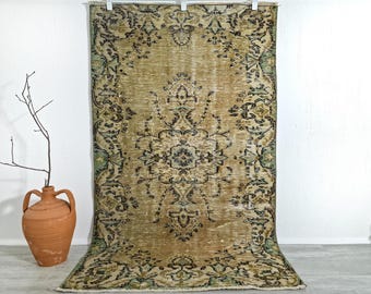 Puede incluir: Una alfombra rectangular con un medallón central y complejos patrones florales en los bordes, en tonos oro apagado, verde oliva y gris carbón oscuro. La alfombra presenta una textura desgastada con flecos visibles en el borde superior.