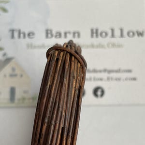 May include: Close-up of a bundle of twisted copper wire, tightly bound at the top. The wire has a weathered, aged appearance with varying shades of brown and copper. The background is a blurred white card with text.