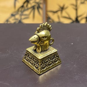 Peut inclure: Petite figurine en forme de tête de coq de couleur laiton, posée sur une base carrée ornée de motifs ajourés. Le coq présente des détails précis, notamment une crête, un bec et un œil. La base est légèrement inclinée.