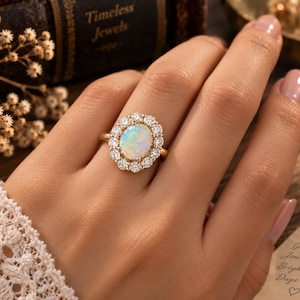 May include: A gold ring featuring a central oval-shaped iridescent opal gemstone surrounded by a halo of small round clear stones. The ring band is thin and polished gold, showcasing a classic design with a prominent, multi-colored gemstone center.