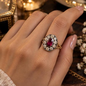 May include: A gold ring featuring a central oval-cut deep red ruby surrounded by a halo of ten round-cut white diamonds. The diamonds are set in a dark metal frame, while the band is crafted from polished yellow gold with a split shank design.