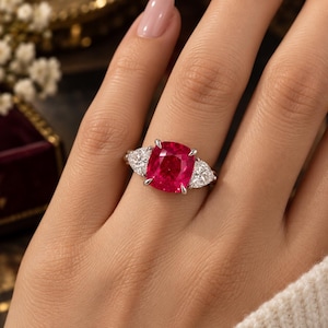 Puede incluir: Un anillo de tono plateado con una gran piedra preciosa de color rojo vibrante en talla cojín en el centro, flanqueada por dos piedras blancas transparentes más pequeñas en forma de pera, montado en una banda de metal pulido con cuatro garras.