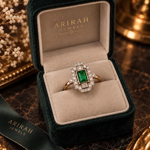 May include: A gold ring featuring a rectangular emerald center stone surrounded by a geometric halo of small round diamonds. The ring is displayed inside a dark green velvet box with the text ARIRAH JEWELS printed in gold on the inner lid.