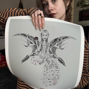 May include: A black and white illustration of a fairy with large, detailed wings, holding a red object. The fairy is wearing a corset and has long hair. The artwork has a gothic theme and includes handwritten text at the bottom.
